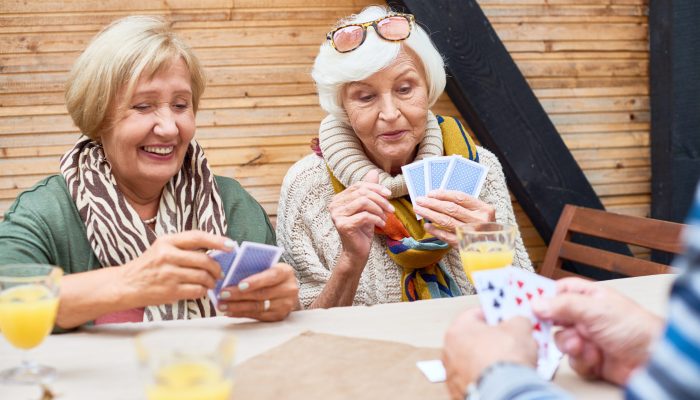 Joyful senior women wearing warm clothes playing poker with friends while sitting at outdoor cafe table